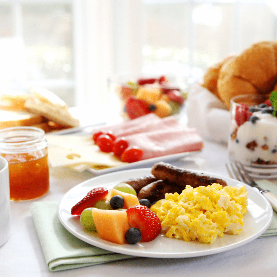 Breakfast spread with scrambled eggs, sausages, fruit, yogurt, ham, cheese, croissants, and coffee on a sunlit table.