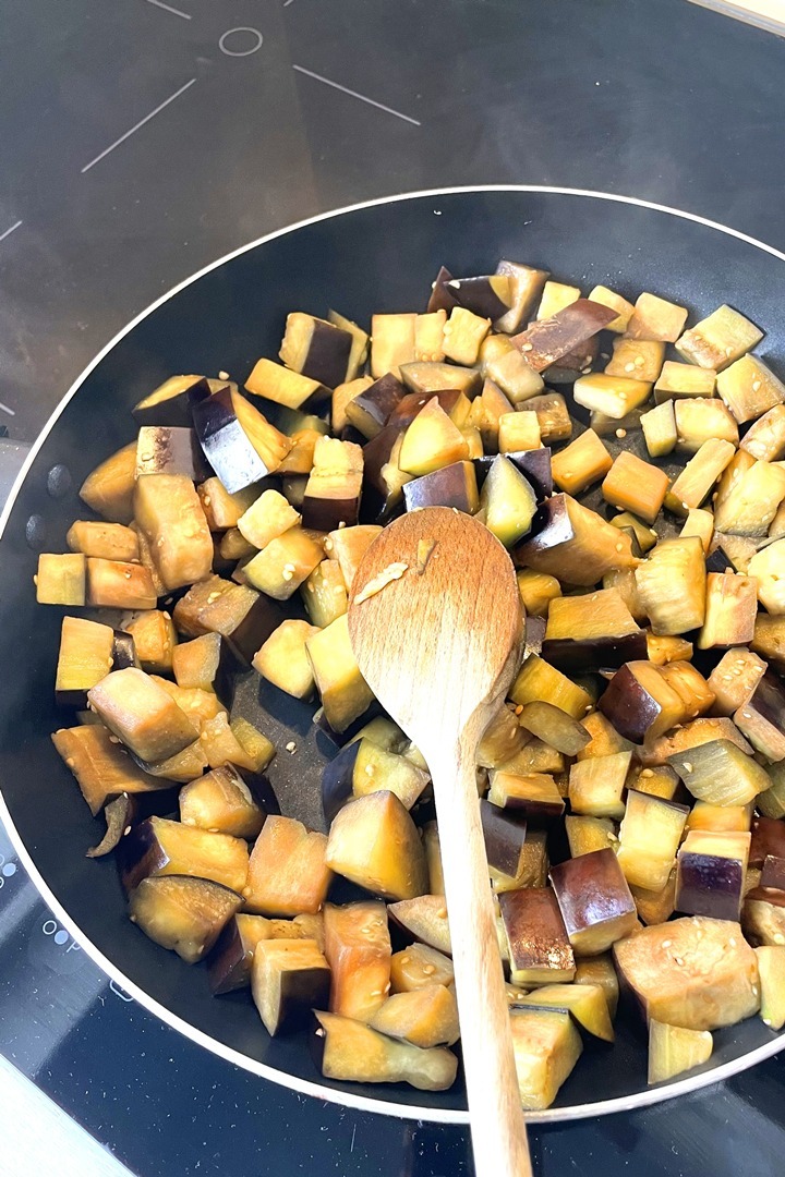 Diced eggplant being sautéed in a pan with a wooden spoon on a stovetop.