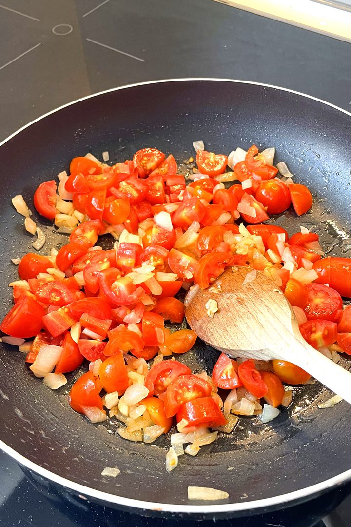 Chopped tomatoes and onions being sautéed in a skillet with a wooden spoon.