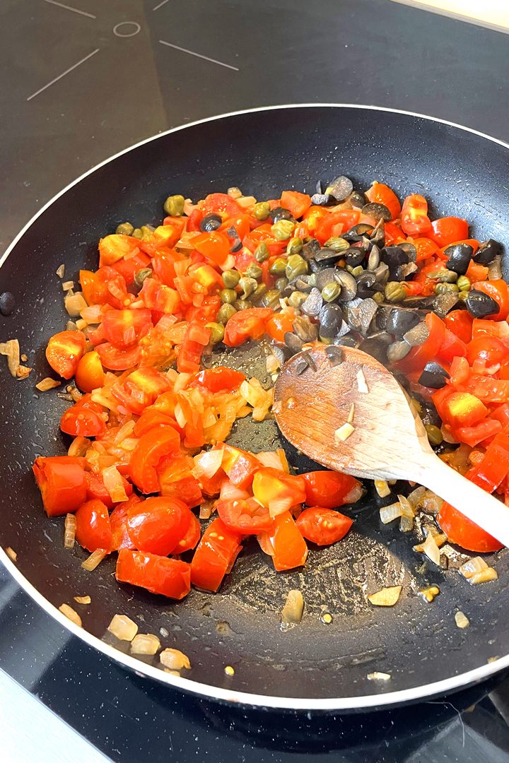 Chopped tomatoes, olives, and capers sautéing in a pan with a wooden spoon on a stovetop.