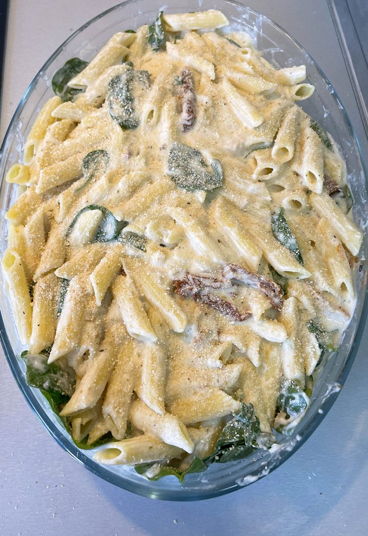 Creamy baked penne pasta with spinach and sun-dried tomatoes, topped with cheese in a glass dish.