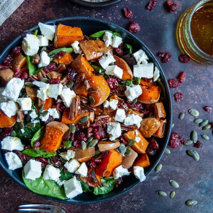 Colorful autumn salad with roasted butternut squash, feta cheese, pecans, and cranberries on a dark rustic table.
