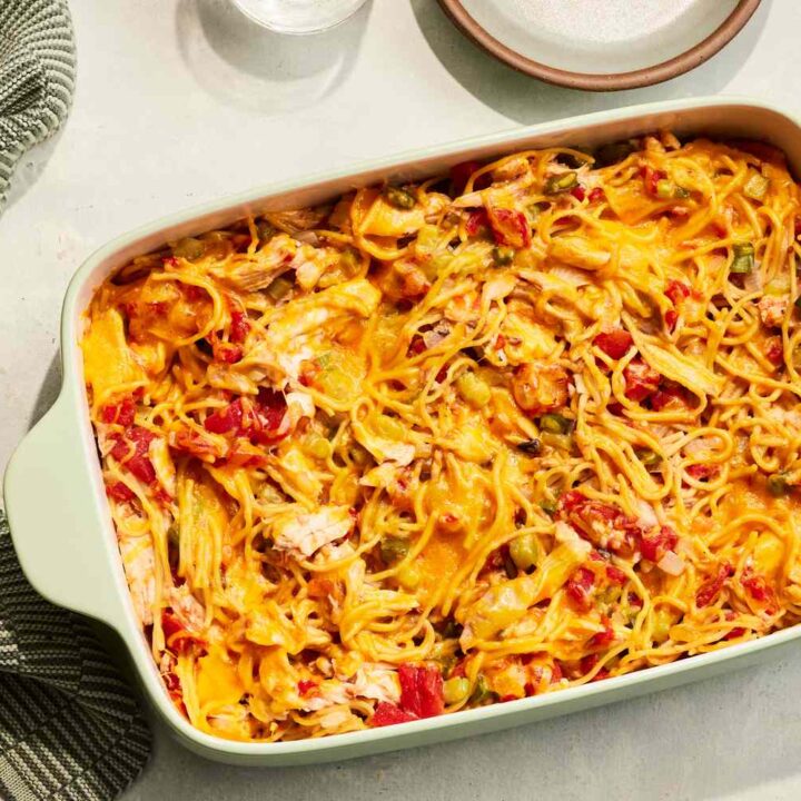 Cheesy chicken spaghetti casserole in a green baking dish, placed on a table with plates and a spoon nearby.