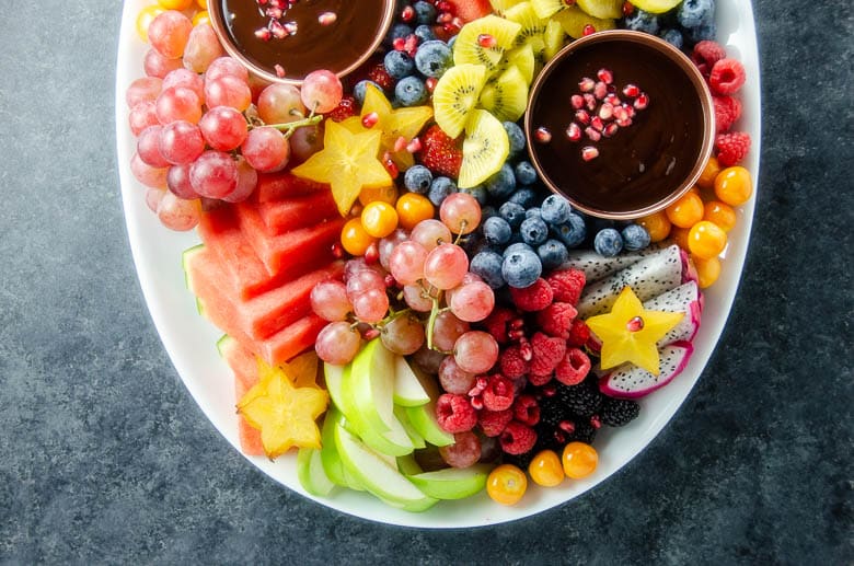 Colorful fruit platter with grapes, kiwi, berries, starfruit, and chocolate dip on a white plate. Perfect for parties.