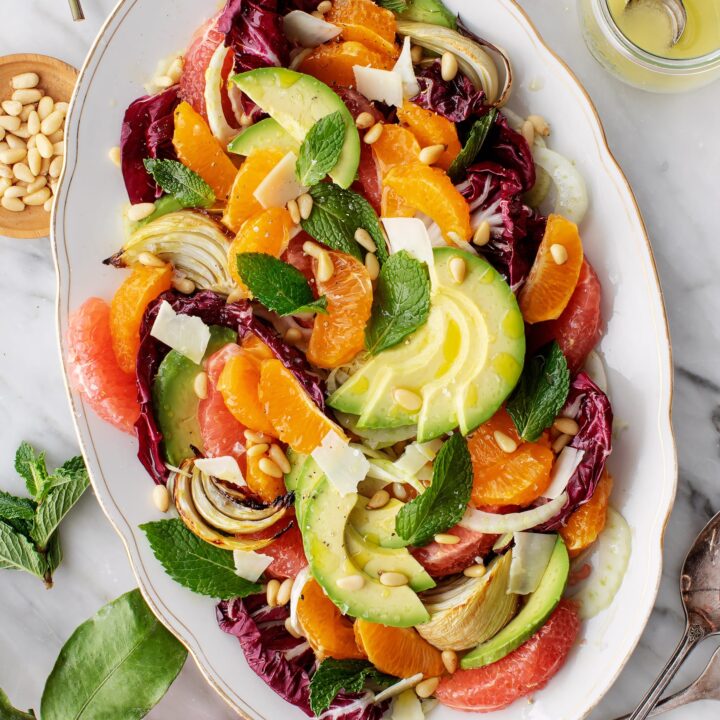Vibrant citrus avocado salad with pine nuts, mint, and shaved cheese on a white platter. Fresh and colorful dish.