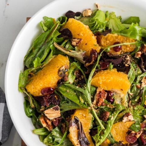 Fresh green salad with orange slices, dried cranberries, and nuts in a white bowl on a marble surface.