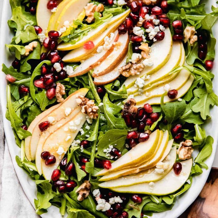 Fresh arugula salad with sliced pears, pomegranate seeds, walnuts, and feta cheese on a white platter.