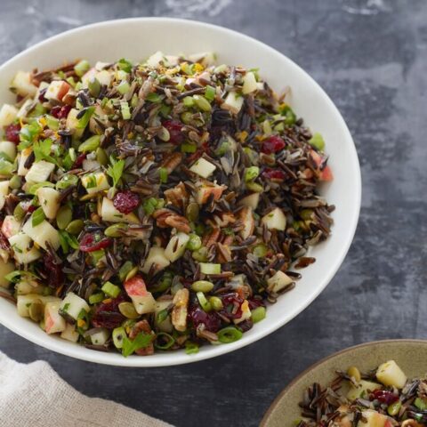 Bowl of vibrant wild rice salad with apples, nuts, and cranberries on a textured background.