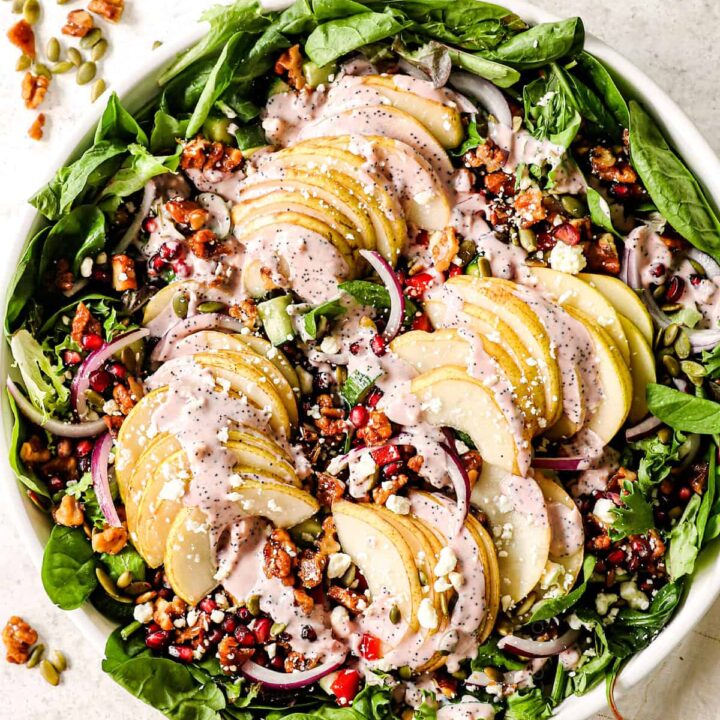 Fresh apple and spinach salad with walnuts, feta, and creamy dressing, garnished with seeds and onions in a white bowl.