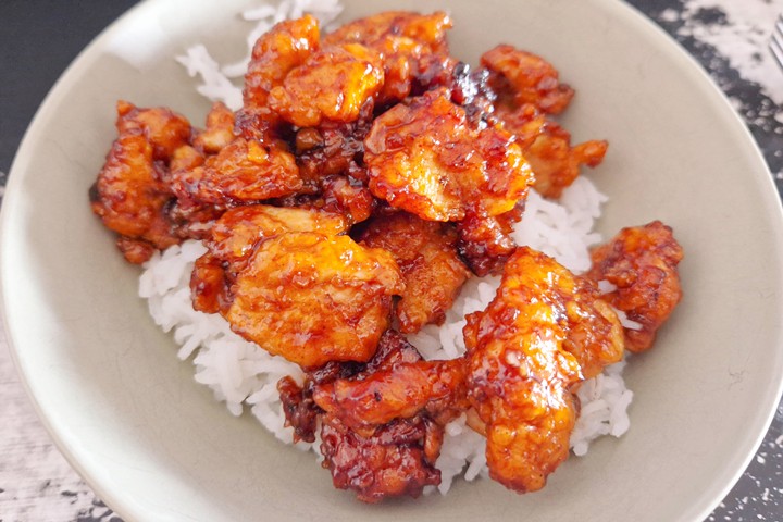 Crispy chicken coated in sweet sauce served over a bed of fluffy white rice in a bowl. Perfect for a delicious meal.