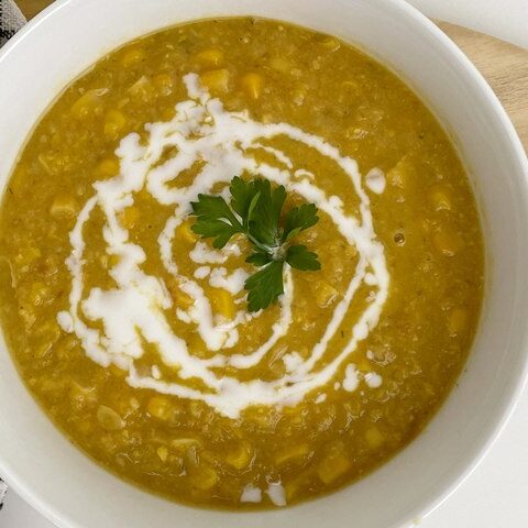 Bowl of homemade creamy corn soup garnished with parsley and a swirl of cream, served on a wooden board with a napkin.