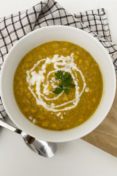 Bowl of creamy corn soup garnished with fresh parsley and cream swirl, served on a checkered cloth.
