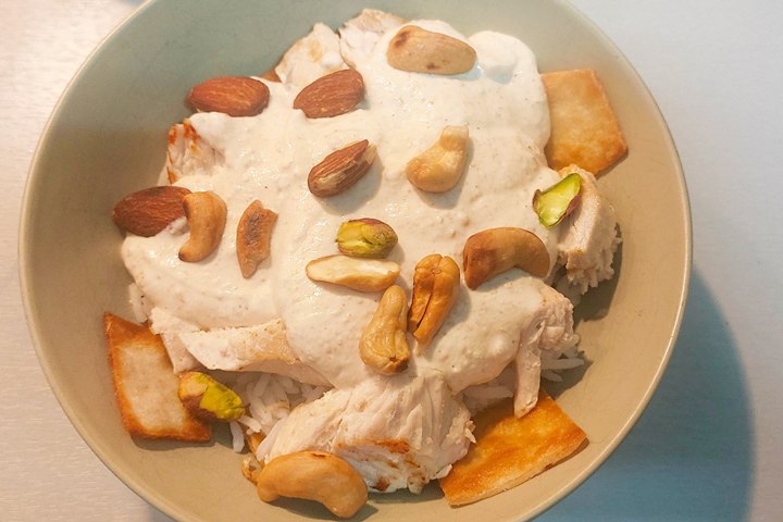 Bowl of chicken with creamy sauce, mixed nuts, rice, and bread pieces, served in a light green dish.