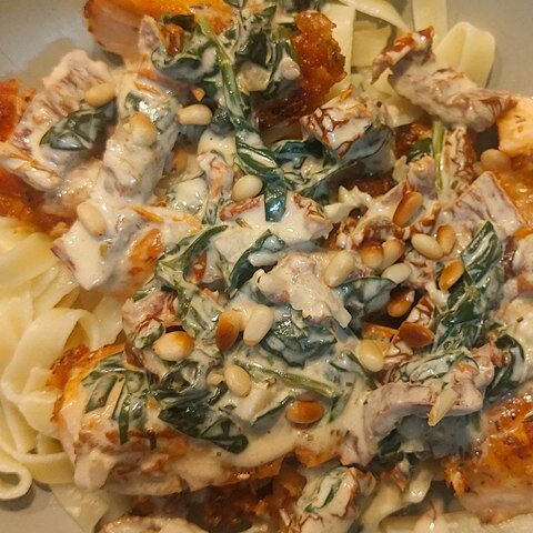 Creamy pasta with chicken, sun-dried tomatoes, spinach, and pine nuts in a rich sauce on a gray plate.