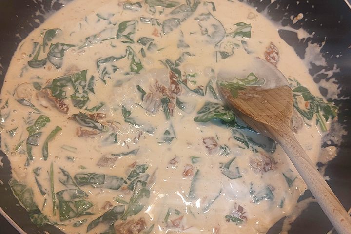 Creamy pasta sauce with spinach and sun-dried tomatoes stirred with a wooden spoon in a pan.