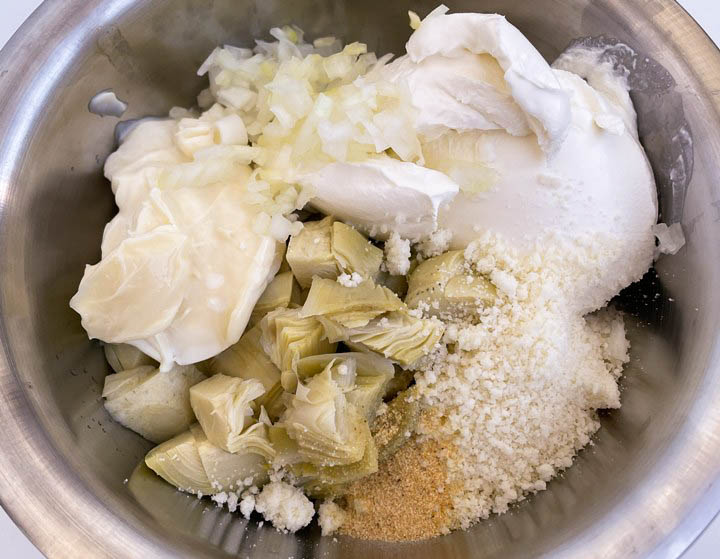 Creamy artichoke dip ingredients in a metal bowl: artichokes, sour cream, onion, cheese, and seasonings. Perfect for parties!
