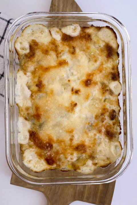 Golden-brown baked artichoke dip in glass dish, topped with melted cheese, resting on a wooden cutting board.
