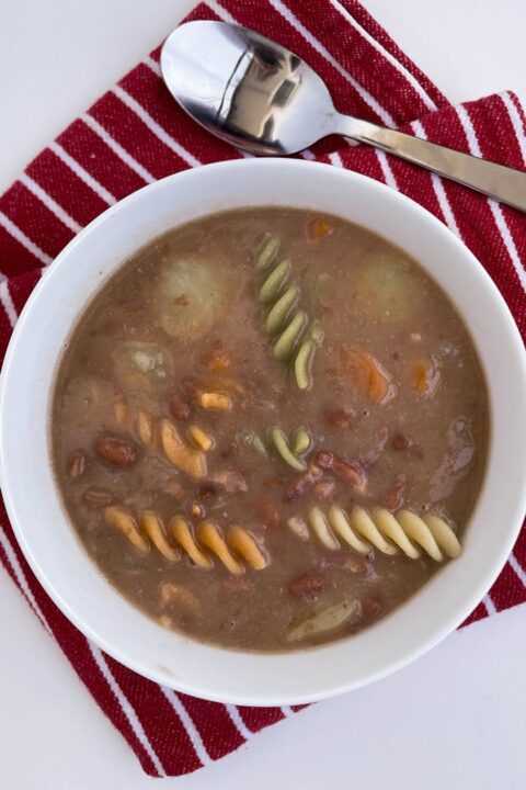 Delicious bowl of hearty soup with colorful pasta and beans, resting on a red striped cloth with a spoon.