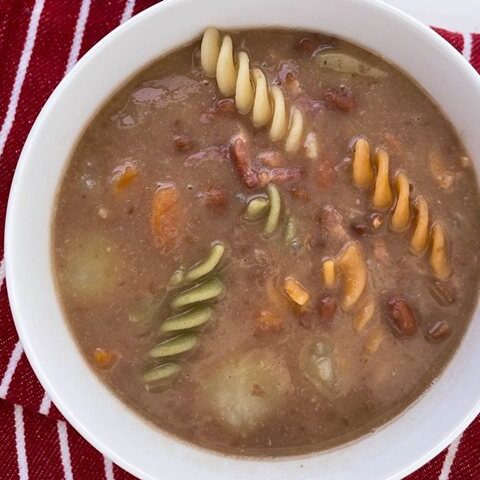 Hearty soup with colorful pasta, beans, and vegetables in a white bowl on a striped red napkin. Perfect comfort food!