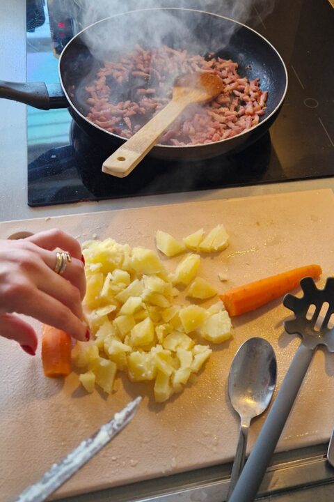 Sautéing bacon in a pan while chopping potatoes and carrots on a cutting board in a kitchen. Steam rising from the pan.