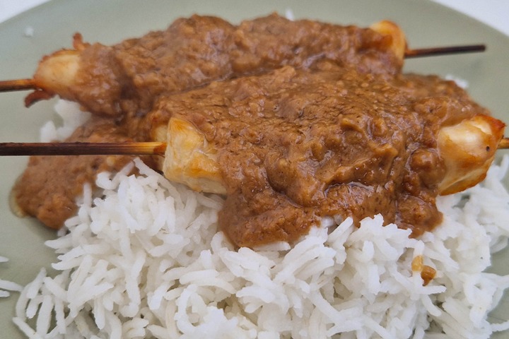 Delicious chicken skewers topped with peanut sauce, served over fluffy white rice on a light green plate.