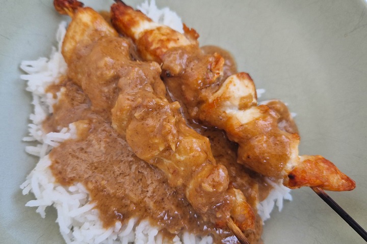 Grilled chicken skewers drizzled with rich peanut sauce on a bed of white rice, served in a green bowl.