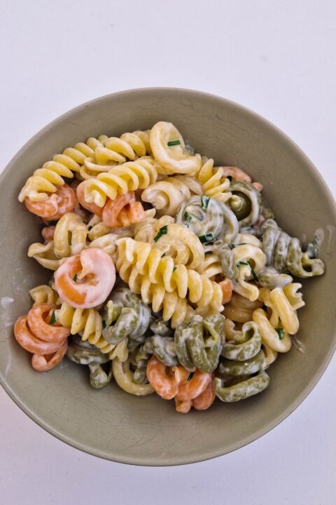 Colorful pasta salad with yellow, green, and orange fusilli in a creamy sauce, garnished with herbs in a bowl.