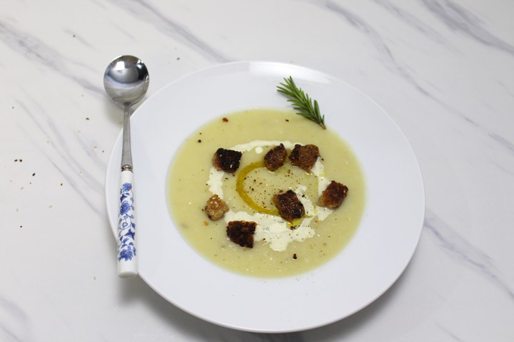 Creamy soup with croutons, drizzled with olive oil and a sprig of rosemary, served in a white bowl on a marble surface.